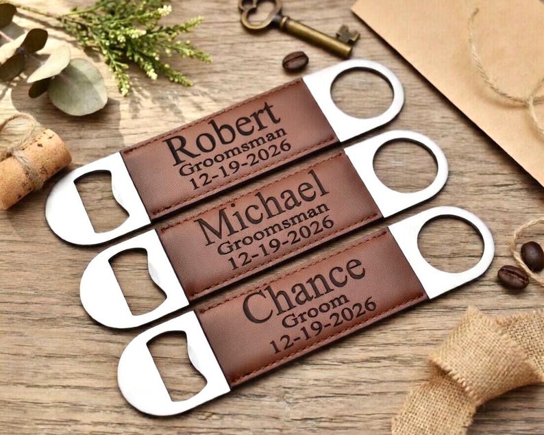May include: Three personalized bottle openers with brown leather and silver metal. Each opener has a name and date engraved: Robert, Michael, and Chance. The text reads "Groomsman" and "Groom" respectively. A perfect gift for a wedding party.
