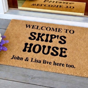 May include: A rectangular doormat made of natural coir fiber. The mat features the words "WELCOME TO SKIP'S HOUSE" in bold black letters, with "John & Lisa live here too." below. The mat is placed on a gray wooden porch.