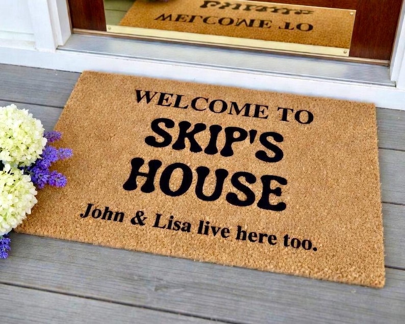 May include: A rectangular doormat made of natural coir fiber. The mat features the words "WELCOME TO SKIP'S HOUSE" in bold black letters, with "John & Lisa live here too." below. The mat is placed on a gray wooden porch.