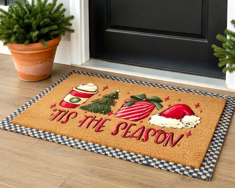 May include: A rectangular doormat with a tan background and a blue and white checkered border. The mat features festive illustrations of a coffee cup, Christmas tree, wrapped gift, and Santa hat, with the words "'Tis the Season" in red.