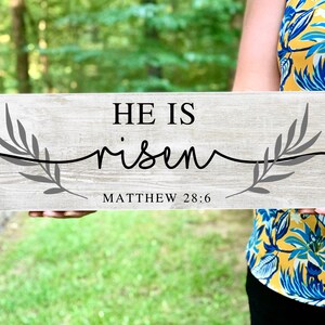 May include: A wooden sign with a gray wash finish, featuring the text "HE IS risen" in black lettering with a gray branch design on either side. The text "MATTHEW 28:6" is printed below the main text.
