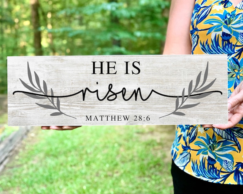 May include: A wooden sign with a gray wash finish, featuring the text "HE IS risen" in black lettering with a gray branch design on either side. The text "MATTHEW 28:6" is printed below the main text.