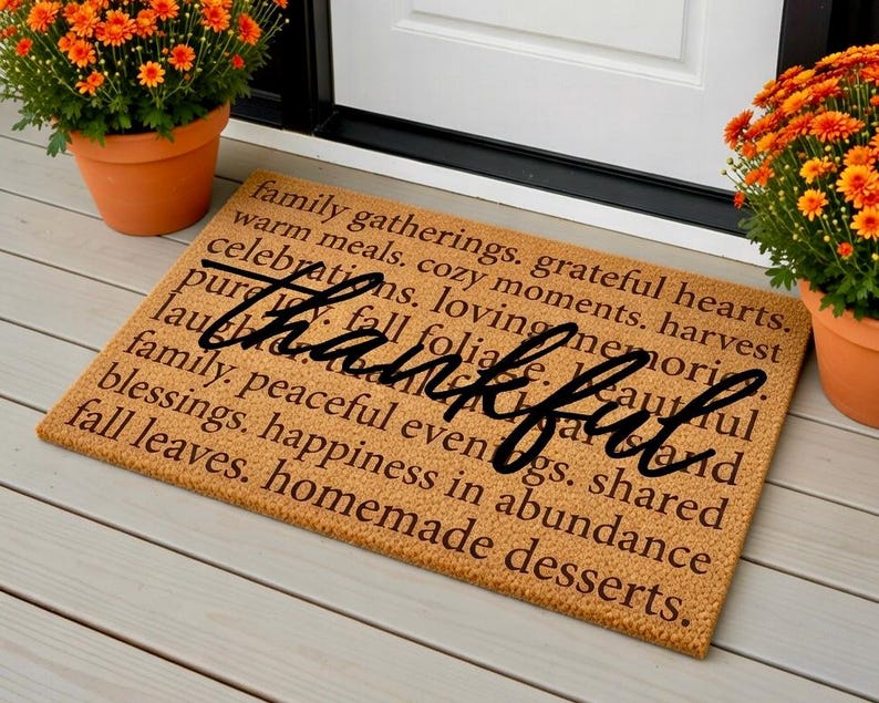 May include: A rectangular doormat with the word "Thankful" in a cursive font, surrounded by words like "family," "grateful," and "harvest." The mat is tan, and the text is dark brown. Orange flowers in terracotta pots flank the door.