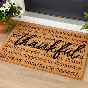 May include: A rectangular doormat with the word "thankful" in a cursive font. The mat is brown with printed text including "family gatherings" and "homemade desserts". Pots of flowers flank the mat.