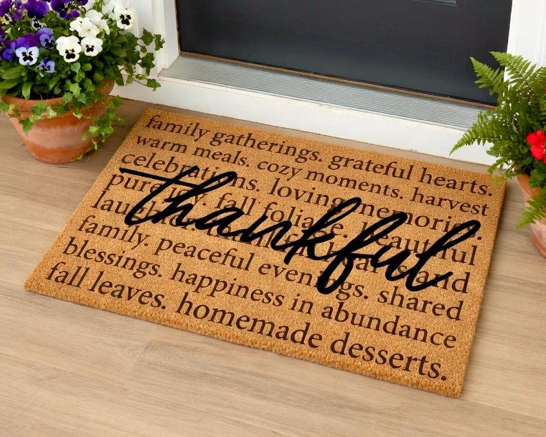 May include: A rectangular doormat with the word "thankful" in a cursive font. The mat is brown with printed text including "family gatherings" and "homemade desserts". Pots of flowers flank the mat.
