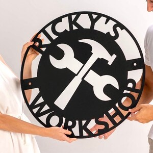 May include: Black metal sign with the text "Ricky's Workshop" in a circular design. A wrench and hammer are crossed in the center of the sign.