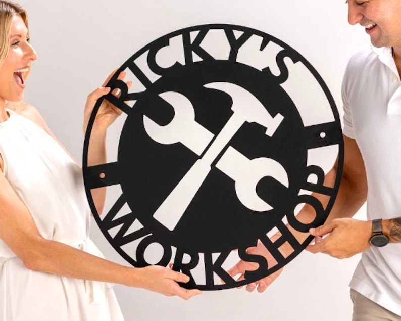 May include: Black metal sign with the text "Ricky's Workshop" in a circular design. A wrench and hammer are crossed in the center of the sign.