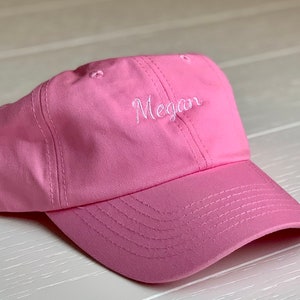 May include: A pink baseball cap with a curved peak. The word "Megan" is embroidered in white script on the front. The cap is made of a soft fabric and has a structured crown.
