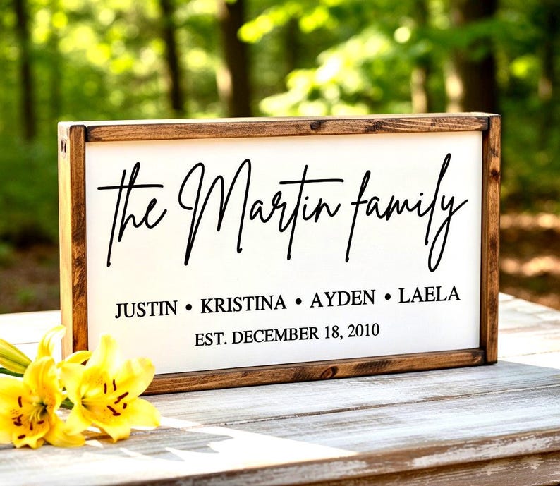 May include: A wooden framed sign with the words "the Martin family" in elegant black script. Below, the names Justin, Kristina, Ayden, and Laela are listed. The date "EST. DECEMBER 18, 2010" is also included. Yellow flowers are in the foreground.