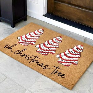 May include: A rectangular doormat with three Christmas tree designs in red and white stripes and polka dots. The text "oh, christmas tree" is written in a cursive font across the mat. The mat is made of a natural fiber material.