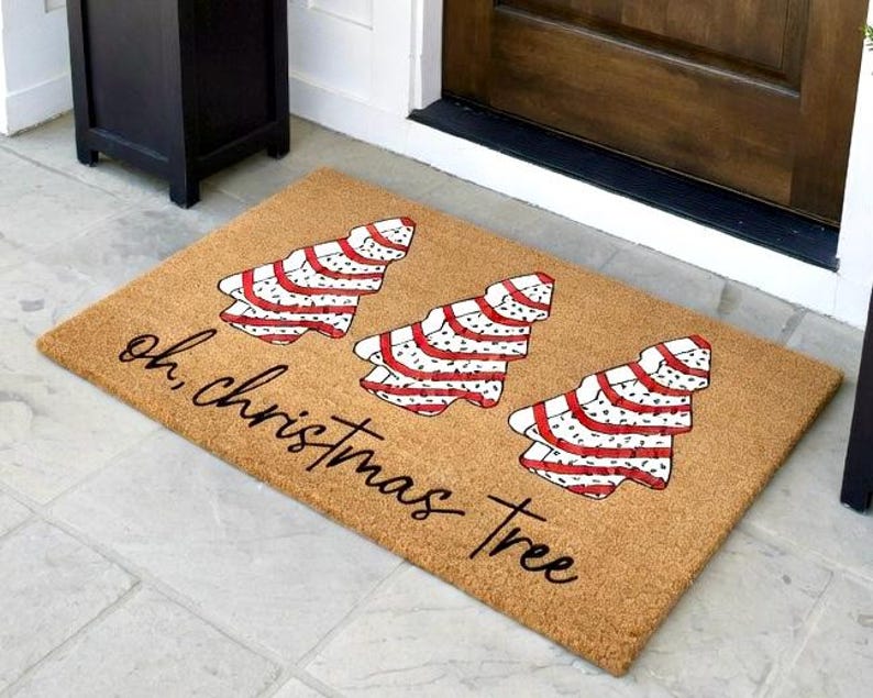 May include: A rectangular doormat with three Christmas tree designs in red and white stripes and polka dots. The text "oh, christmas tree" is written in a cursive font across the mat. The mat is made of a natural fiber material.