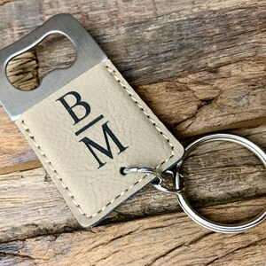 May include: A beige leather bottle opener keychain with a silver bottle opener and a silver key ring. The keychain is engraved with the initials "B|M".