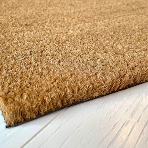 May include: Close-up of a rectangular doormat made of natural coir fibers. The mat has a light brown color and a rough, textured surface. The edge of the mat is dark, and the background is a white wooden surface.