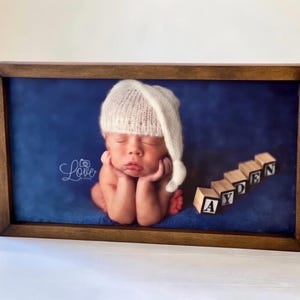 May include: A framed photograph of a newborn baby wearing a white knit cap, resting on a blue backdrop. Wooden blocks spell out the name "AYDEN". The frame is made of dark brown wood. The image has a soft, gentle aesthetic.