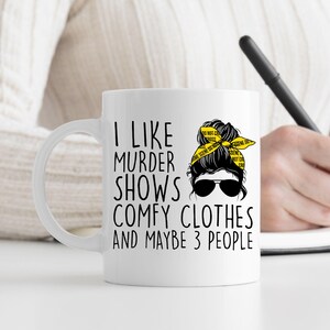 May include: White ceramic mug with a black and yellow design. The mug features a cartoon woman with a messy bun and the text "I like murder shows comfy clothes and maybe 3 people".