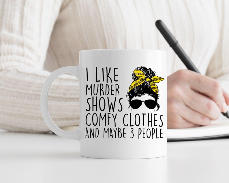 May include: White ceramic mug with a black and yellow design. The mug features a cartoon woman with a messy bun and the text "I like murder shows comfy clothes and maybe 3 people".