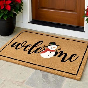 K&ouml;nnte beinhalten: Eine rechteckige Fu&szlig;matte mit dem Wort "welcome" in schwarzer Schrift und einer Schneemann-Grafik. Die Matte ist beige mit einem schwarzen Rand. Topfrote Weihnachtsstern flankieren eine braune T&uuml;r.