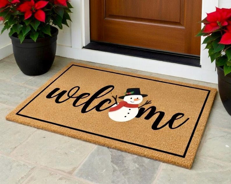 K&ouml;nnte beinhalten: Eine rechteckige Fu&szlig;matte mit dem Wort "welcome" in schwarzer Schrift und einer Schneemann-Grafik. Die Matte ist beige mit einem schwarzen Rand. Topfrote Weihnachtsstern flankieren eine braune T&uuml;r.