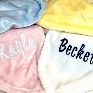 May include: Four plush blankets with embroidered names. The blankets are in shades of blue, yellow, pink, and white. The names are "Kayla", "Becket", and two others that are not visible.