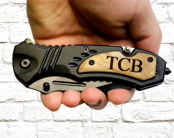 May include: Black and silver folding pocket knife with a wooden handle. The handle is engraved with the initials "TCB". The knife is held in a hand.