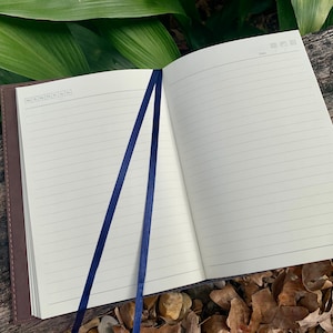 May include: Open notebook with lined pages and a dark brown cover. Two navy blue ribbon bookmarks are visible. The notebook is open on a wooden surface with green leaves in the background.