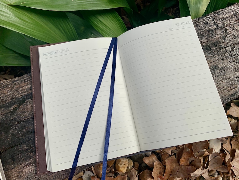 May include: Open notebook with lined pages and a dark brown cover. Two navy blue ribbon bookmarks are visible. The notebook is open on a wooden surface with green leaves in the background.
