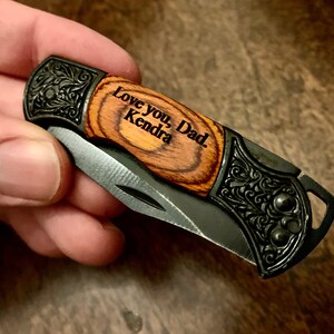 May include: A small pocket knife with a wood handle and a black metal blade. The handle is engraved with the words "Love you, Dad. Kendra".