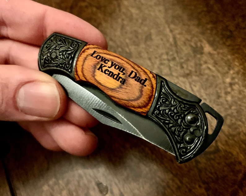 May include: A small pocket knife with a wood handle and a black metal blade. The handle is engraved with the words "Love you, Dad. Kendra".