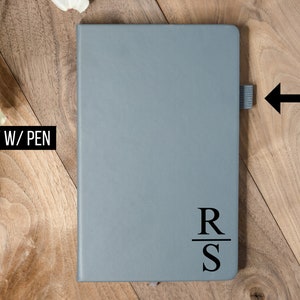 May include: A gray notebook with a pen loop and the initials "RS" printed in black. The notebook is closed and has a simple, minimalist design. The text "GRAY W/ PEN" is in the upper left corner.