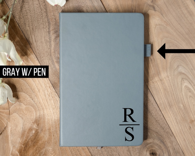 May include: A gray notebook with a pen loop and the initials "RS" printed in black. The notebook is closed and has a simple, minimalist design. The text "GRAY W/ PEN" is in the upper left corner.