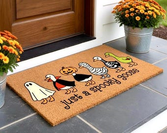 May include: A rectangular doormat with a Halloween theme. The mat features cartoon geese dressed as a ghost, vampire, pumpkin, mummy, and skeleton. The text "just a spooky goose" is printed in black on the mat.