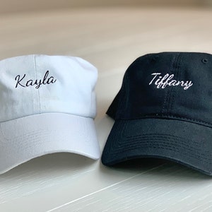 May include: Two baseball caps are shown, one white and one black. The white cap is embroidered with the name "Kayla" in cursive. The black cap has "Tiffany" embroidered on it, also in cursive. Both caps are made of a soft material.
