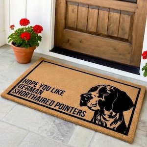 May include: A rectangular doormat with a black border and the text "HOPE YOU LIKE GERMAN SHORTHAIRED POINTERS." A black and white graphic of a German Shorthaired Pointer is on the right side. The mat is tan.