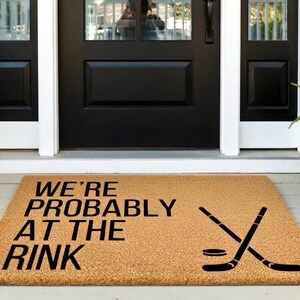 May include: A rectangular doormat made of natural coir fiber. The mat features the text "WE'RE PROBABLY AT THE RINK" in black, along with a graphic of crossed hockey sticks and a puck. Placed in front of a black door.