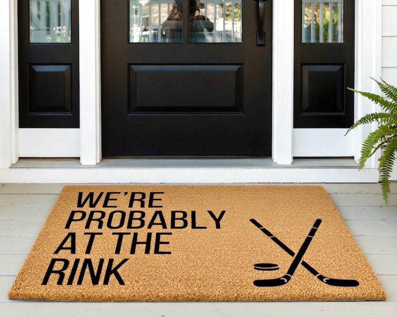 May include: A rectangular doormat made of natural coir fiber. The mat features the text "WE'RE PROBABLY AT THE RINK" in black, along with a graphic of crossed hockey sticks and a puck. Placed in front of a black door.