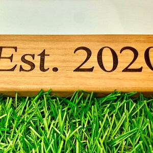 May include: A wooden sign with the text "Est. 2020" engraved in a dark font. The sign is light brown and rests on a bed of bright green artificial grass, with a white background.
