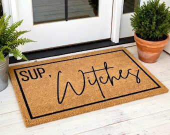 Puede incluir: Una alfombra rectangular hecha de fibra de coco natural. La alfombra tiene un borde negro y presenta las palabras "SUP. Witches" en negro, con una fuente cursiva. La alfombra está colocada frente a una puerta blanca.