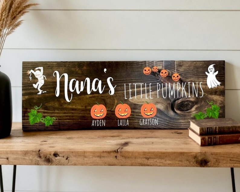 May include: A rustic wooden sign with the text "Nana's Little Pumpkins" in white script. The sign features orange pumpkin and ghost graphics, with the names Ayden, Laila, and Grayson below the pumpkins. A charming Halloween decoration.