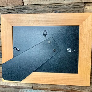 May include: A rectangular wooden picture frame with a light brown finish. The back of the frame is visible, showing a black backing, a hinged easel stand, and metal hanging hardware. The frame is designed to hold a photograph or artwork.