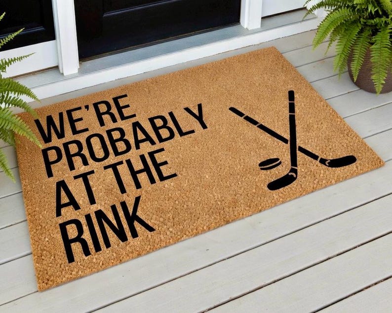 May include: A rectangular doormat made of natural coir fiber. The mat features the text "WE'RE PROBABLY AT THE RINK" in bold black letters, along with crossed hockey sticks and a puck. The mat is placed on a white wooden porch.