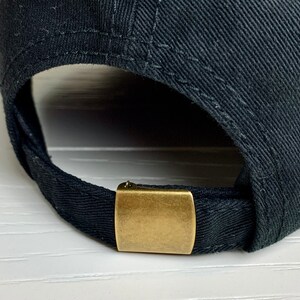 May include: A close-up of a black baseball cap. The cap has a curved peak and an adjustable strap at the back. The strap is secured with a gold-coloured metal clasp.