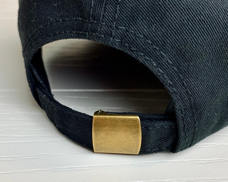 May include: A close-up of a black baseball cap. The cap has a curved peak and an adjustable strap at the back. The strap is secured with a gold-coloured metal clasp.