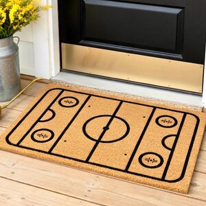 May include: A rectangular doormat with a hockey rink design. The mat is made of a natural tan material with a black hockey rink outline. The rink includes a center circle, face-off circles, and goal creases. Placed in front of a black door.