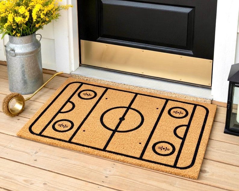 May include: A rectangular doormat with a hockey rink design. The mat is made of a natural tan material with a black hockey rink outline. The rink includes a center circle, face-off circles, and goal creases. Placed in front of a black door.