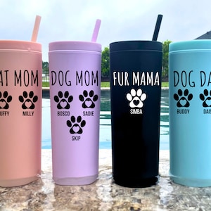 K&ouml;nnte beinhalten: Vier isolierte Becher in Rosa, Lila, Schwarz und Blau. Jeder Becher hat ein Pfotenabdruck-Design mit dem Namen eines Haustiers und einem Titel: "Cat Mom", "Dog Mom", "Fur Mama" und "Dog Dad". Die Becher haben passende Strohhalme.