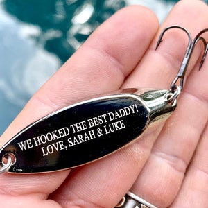 Puede incluir: Se&ntilde;uelo de pesca plateado con una inscripci&oacute;n negra que dice "We hooked the best Daddy! Love, Sarah & Luke"