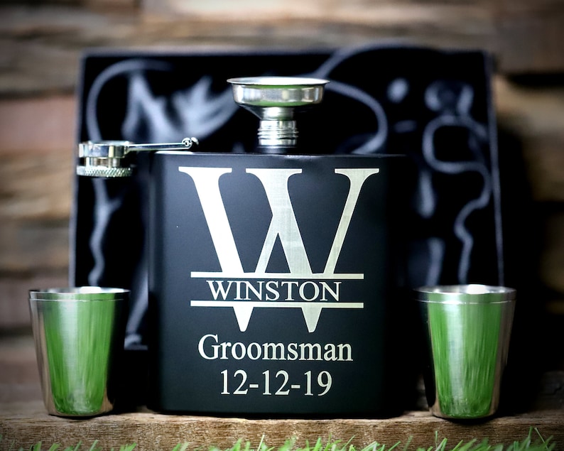 May include: Black flask with a silver funnel and two silver shot glasses. The flask is engraved with a large letter "W" and the text "Winston,  Groomsman, 12-12-19".