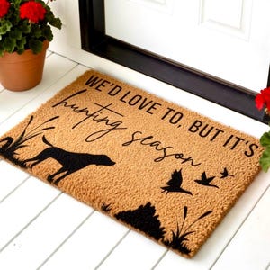 May include: A rectangular doormat with the text "WE'D LOVE TO, BUT IT'S hunting season" in black script. The mat features a black silhouette of a dog, birds, and reeds on a tan background. Decorative potted plants frame the mat.