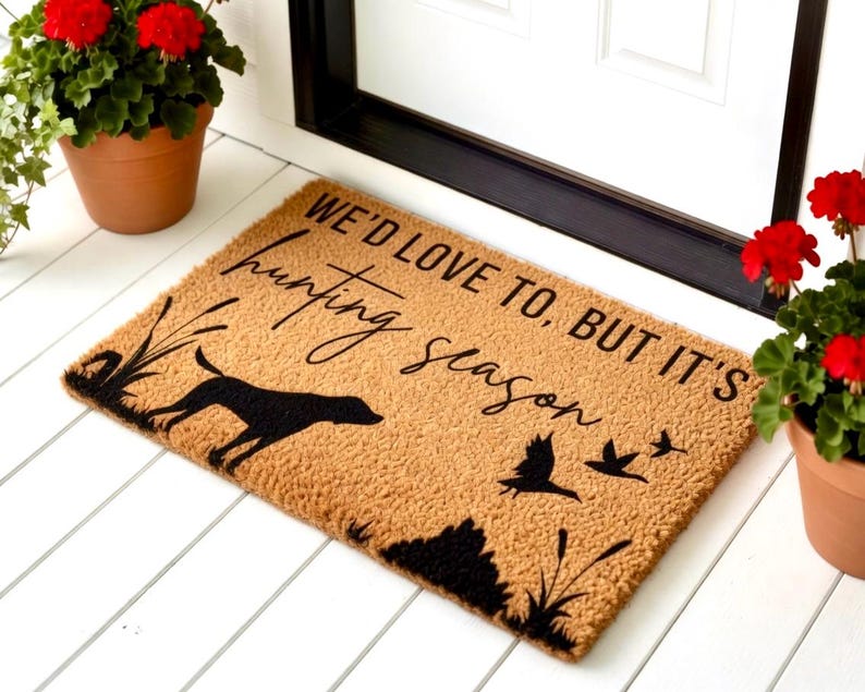 May include: A rectangular doormat with the text "WE'D LOVE TO, BUT IT'S hunting season" in black script. The mat features a black silhouette of a dog, birds, and reeds on a tan background. Decorative potted plants frame the mat.