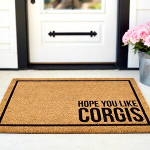 May include: A rectangular doormat made of natural coir fibre, featuring a black border and the text "HOPE YOU LIKE CORGIS" in bold, black letters. The mat is placed in front of a white door.